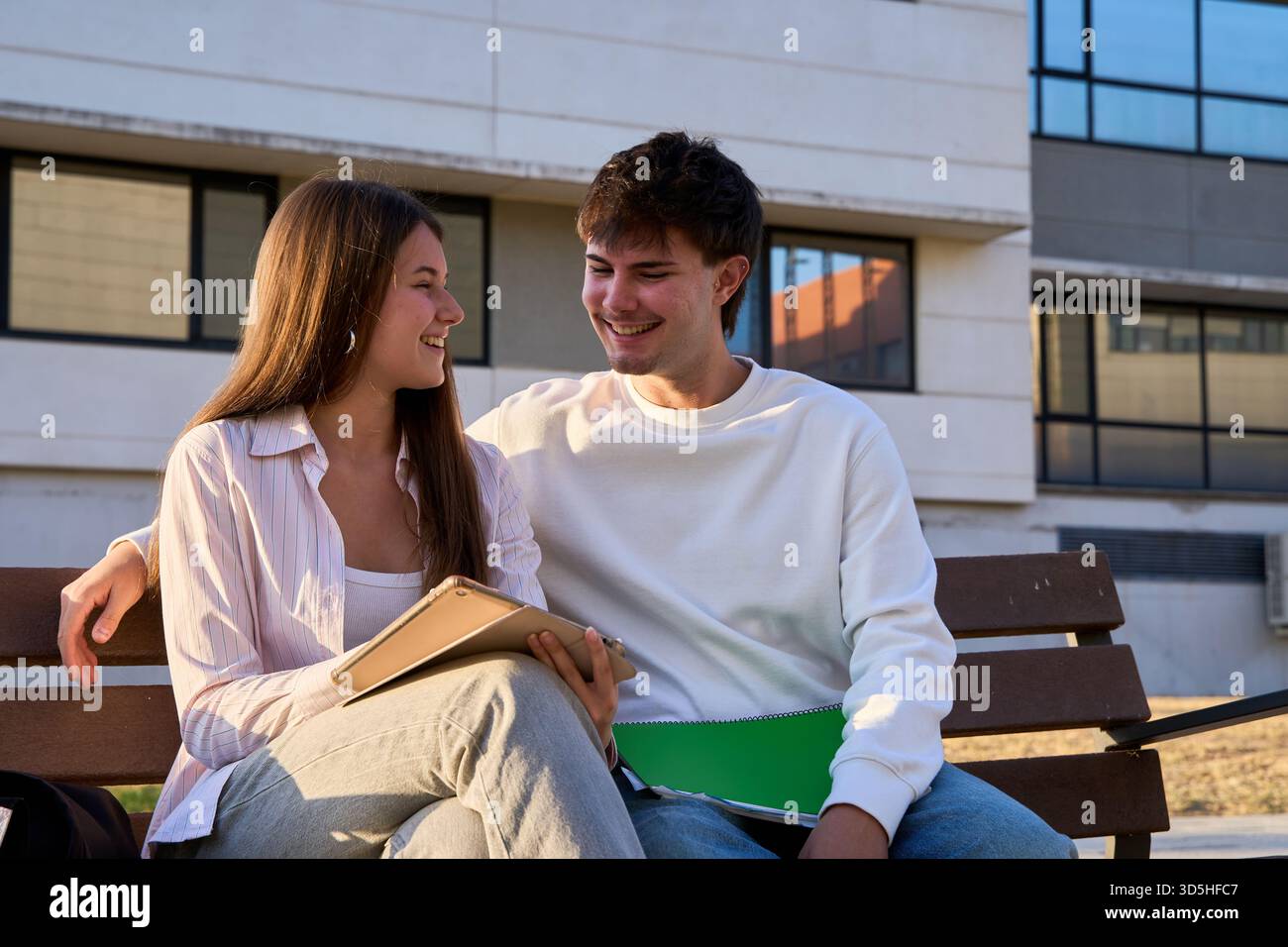 Two cheerful young students sitting on a university campus bench, sharing a moment and looking at a tablet Stock Photo