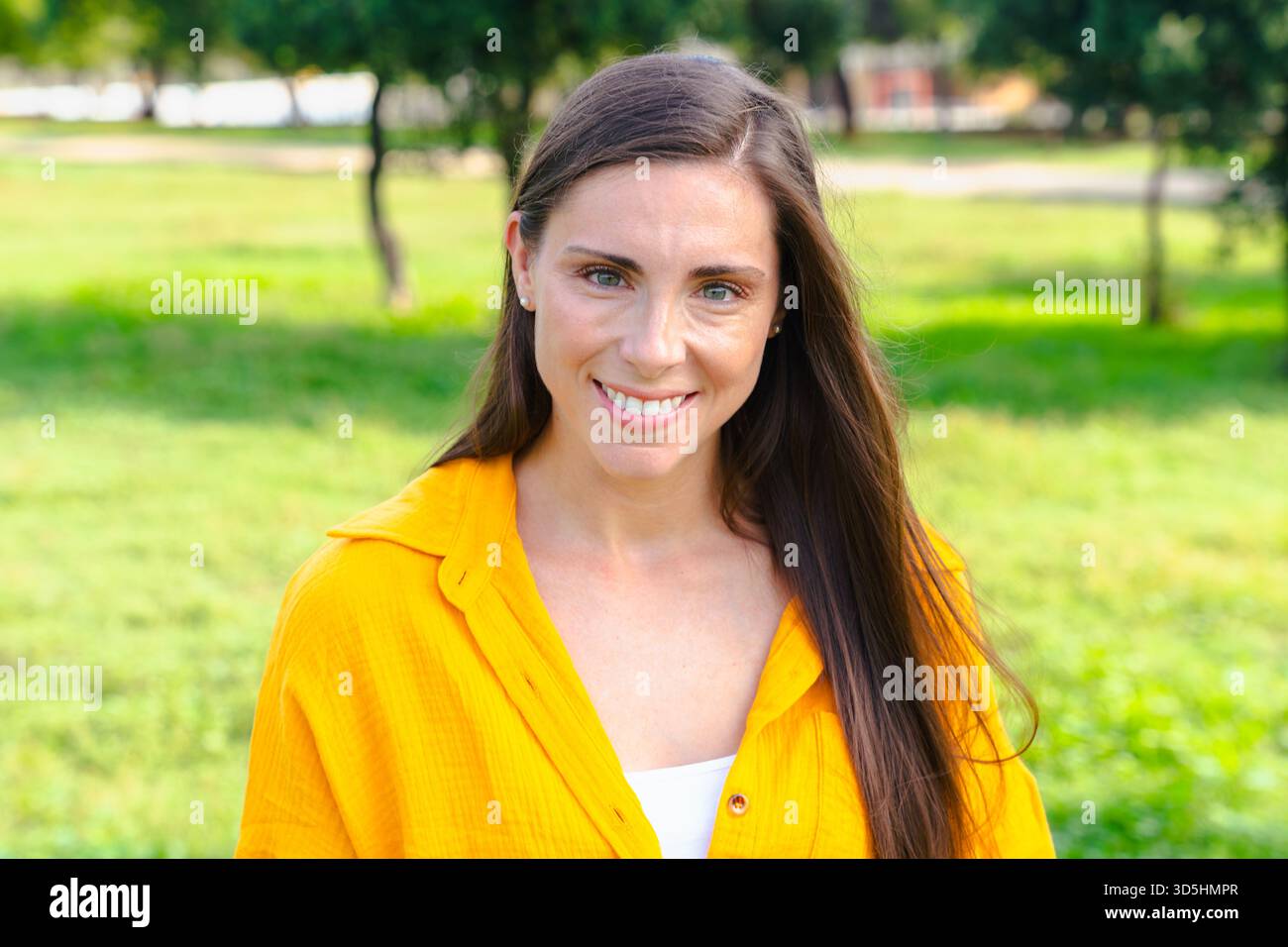 Smiling caucasian woman in yellow shirt looking at camera outdoors in sunny park Stock Photo