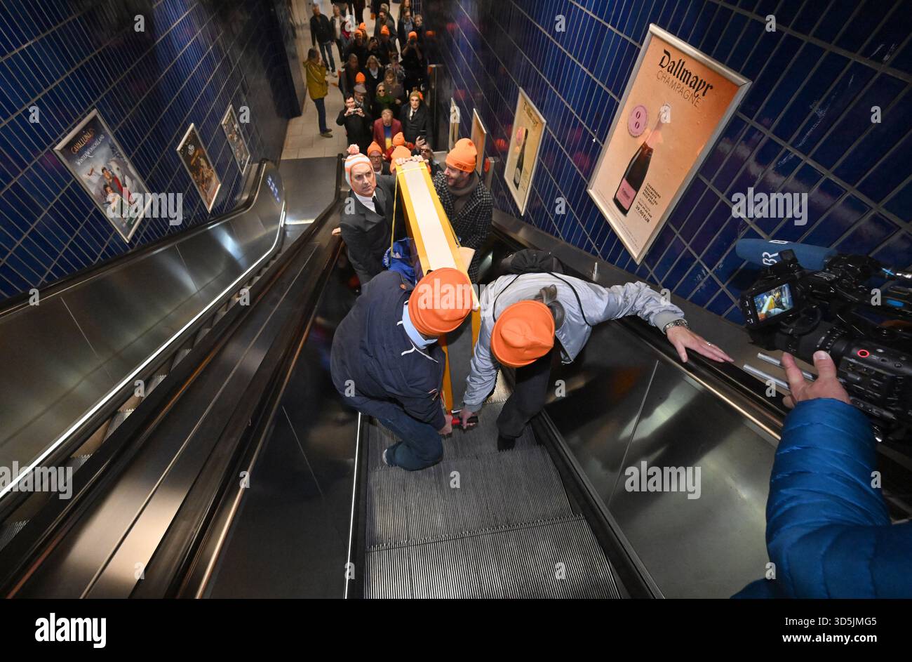 Munich, Germany. 16th Nov, 2025. The Lach- und Schießgesellschaft sign is carried by members of the Münchner Lach- und Schießgesellschaft on a trolley on an escalator to the Marienplatz S-Bahn station. The Münchner Lach- und Schießgesellschaft moves from Haimhauserstraße to the Fat Cat (Alter Gasteig) at Rosenheimer Platz by subway and S-Bahn. Credit: Felix Hörhager/dpa/Alamy Live News Stock Photo