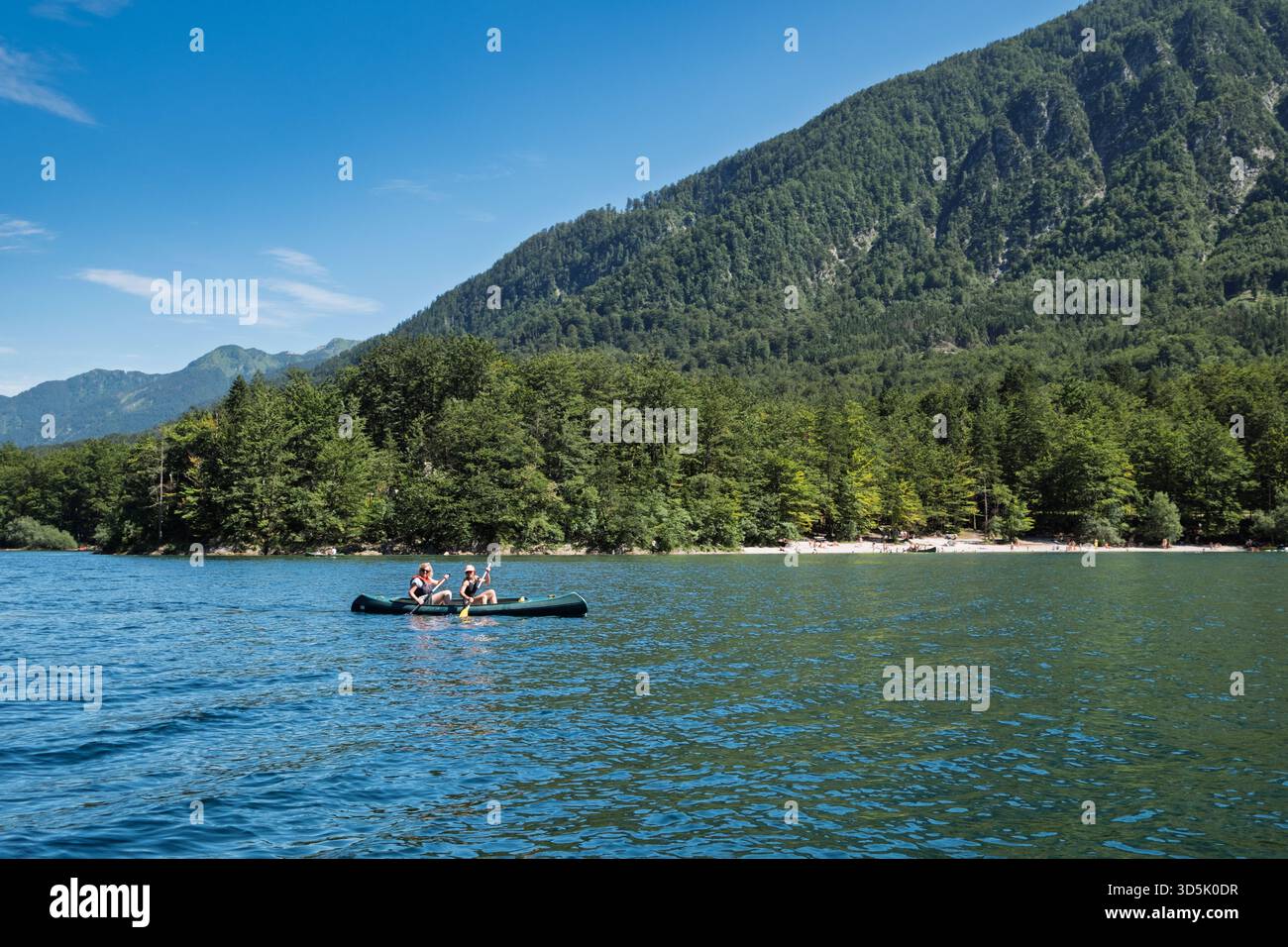 People canoeing on the clear blue waters of Lake Bohinj, Slovenia, with forested mountains and a bright summer sky in the background. Stock Photo