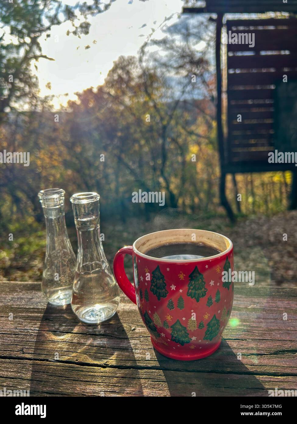 A steaming cup of coffee and two small rakija bottles on a rustic wooden table, illuminated by warm sunlight with a forest backdrop. - Smartphone Captured Stock Image
