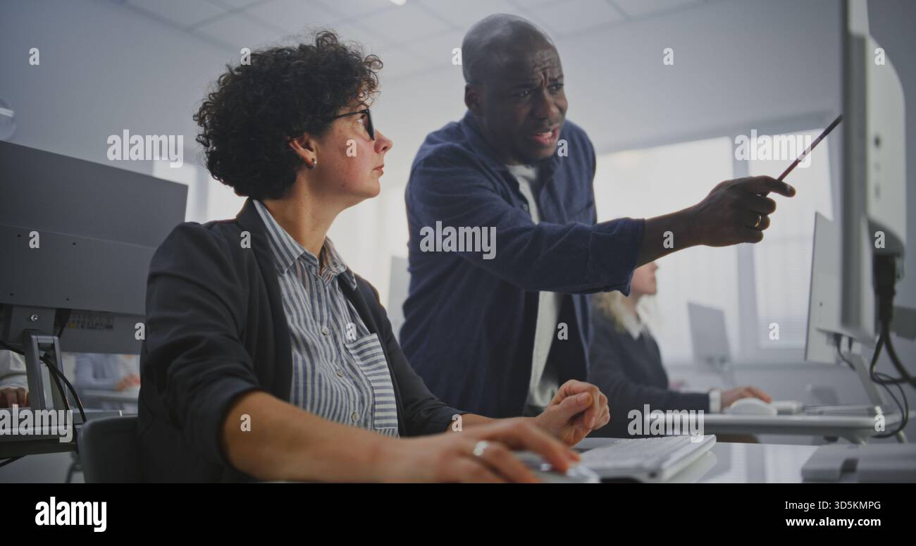 Attentive African American Instructor Walks Between Adult Students During Programming Workshop in Modern Computer Classroom. Helps Debug Code, Provides Feedback, Explains Complex Concepts. Dolly Shot. Stock Photo