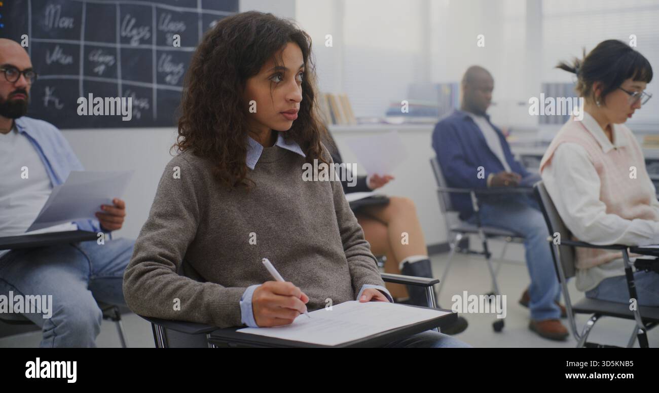 Young Woman in Good Mood Among Group Adult Students of Various Ages Completing Written Test. Students Sitting at Individual Desks and Writing Exam. Concept Professional Training and Lifelong Learning. Stock Photo