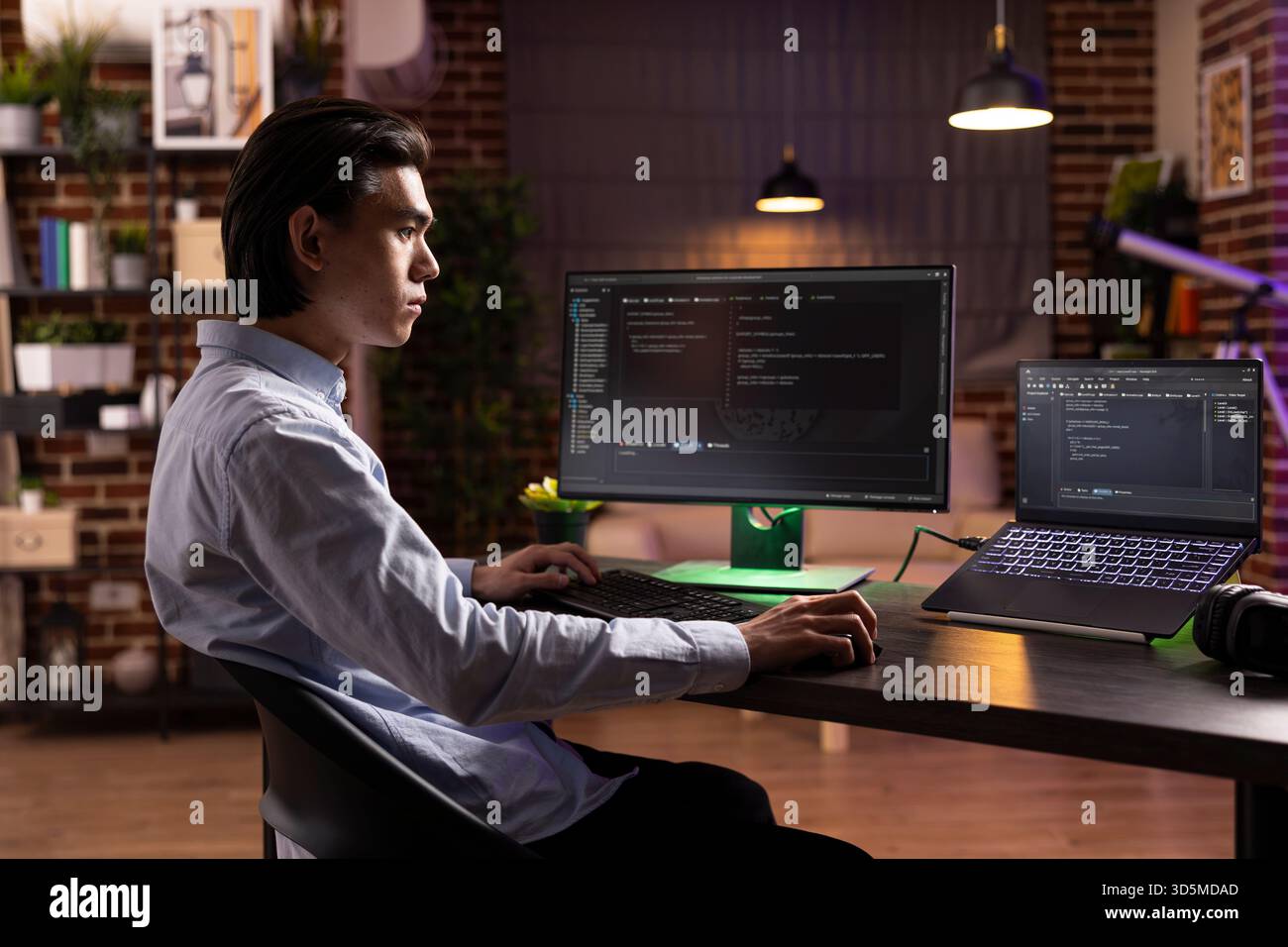 Focused asian man working late, using desktop pc and laptop to debug software. Dedicated remote developer analyzing website code on multiple screens and completing programming tasks at home Stock Photo