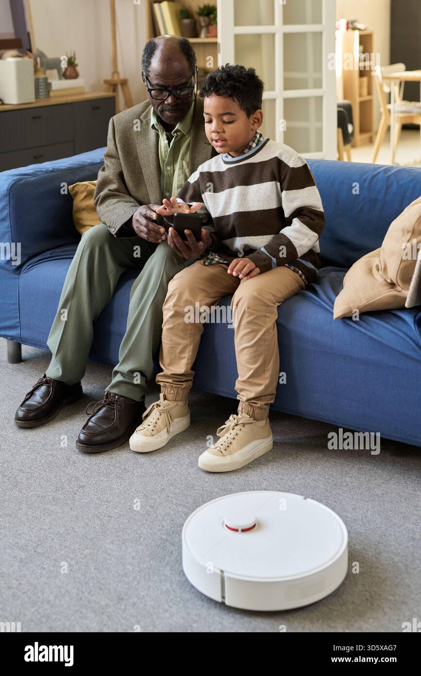 Senior Black Man Teaching Black Boy Using Tablet with Robot Vacuum Stock Photo