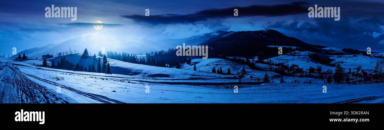 forested valley near village at night. landscape panorama with mountain ridge in winter in full moon light. snow covered rural fields and rolling hill Stock Photo