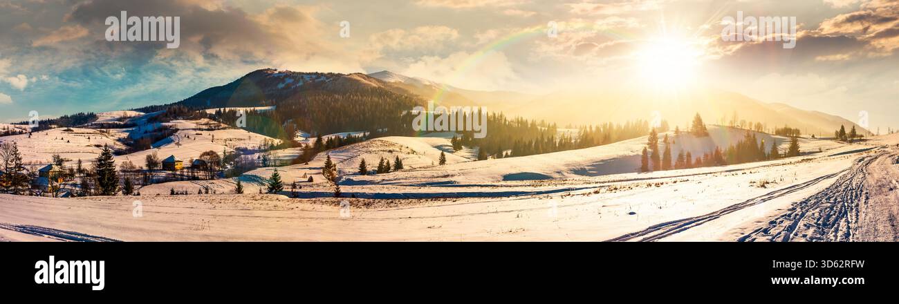 forested valley near village at sunset. landscape panorama with mountain ridge in winter in evening light. snow covered rural fields and rolling hills Stock Photo