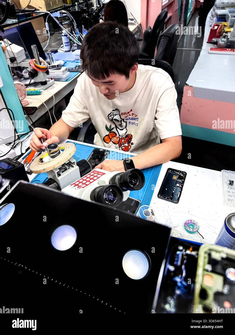 Young asian technician working on electronic components and phones at a cluttered workbench in a tech repair workshop malaysia - Smartphone Captured Stock Image