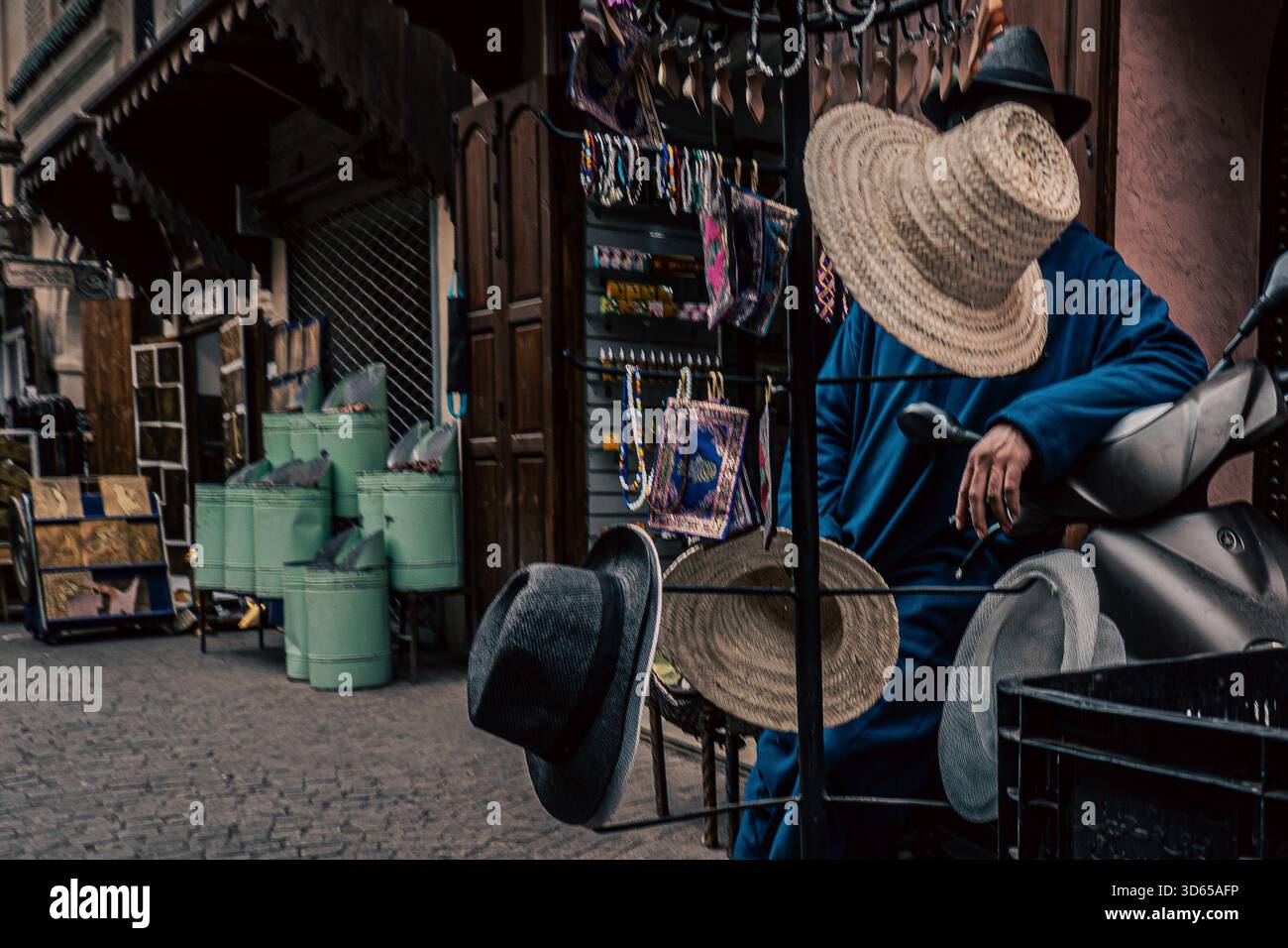 Traditional Moroccan street vendor in Marrakech, selling hats in a narrow street with traditional buildigs. his face is hidden by a hat. Stock Photo