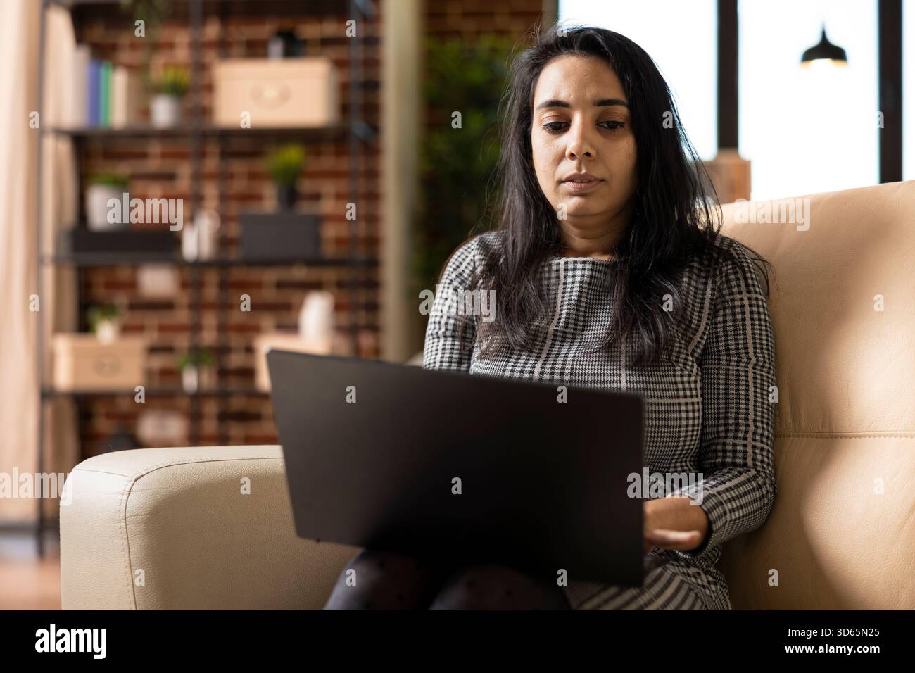 Indian businesswoman seated on cozy sofa, working remotely on laptop, reviewing financial reports and plans. Focused female freelancer using digital device, reading online articles in brick wall home. Stock Photo