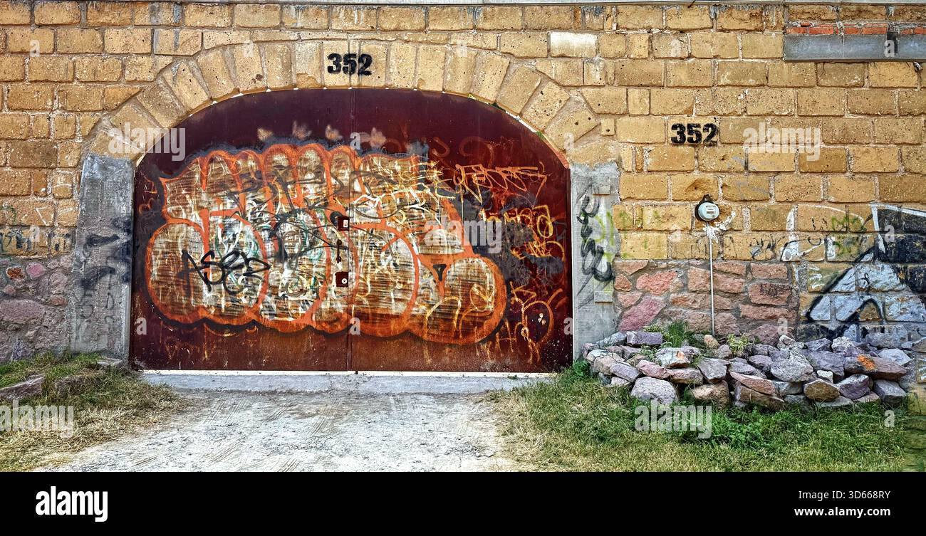 Old rusted metal garage door with colorful graffiti on a stone brick wall in an urban setting with worn surroundings - Smartphone Captured Stock Image