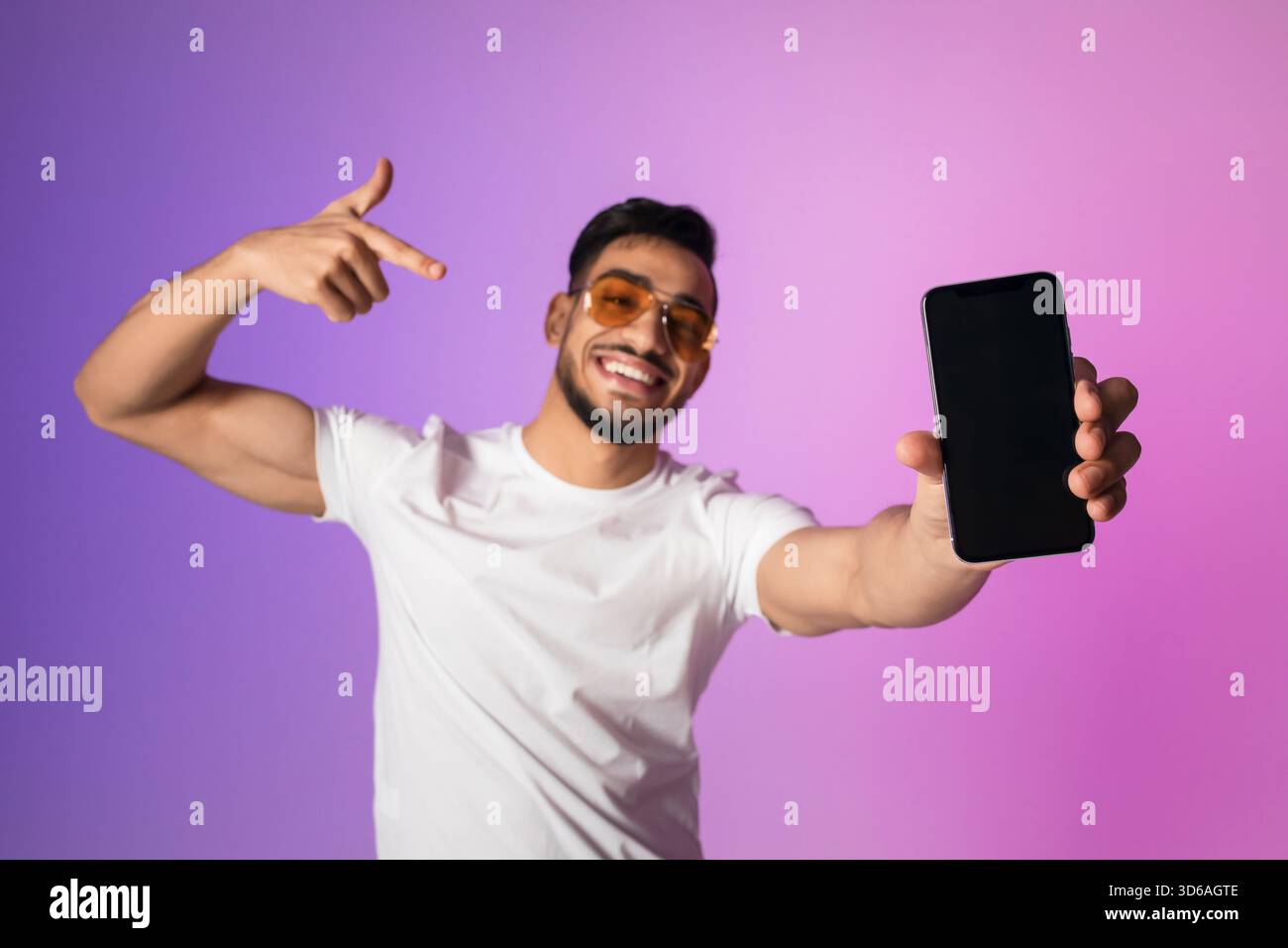 Young Arab Man Smiles and Points at Empty Smartphone Screen in Vibrant Neon Light Setting Stock Photo