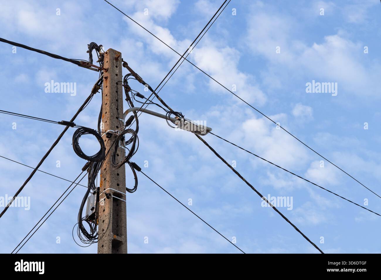 A concrete utility pole featuring a modern LED streetlight and a messy tangle of coiled black cables against a blue sky with fluffy white clouds. Stock Photo
