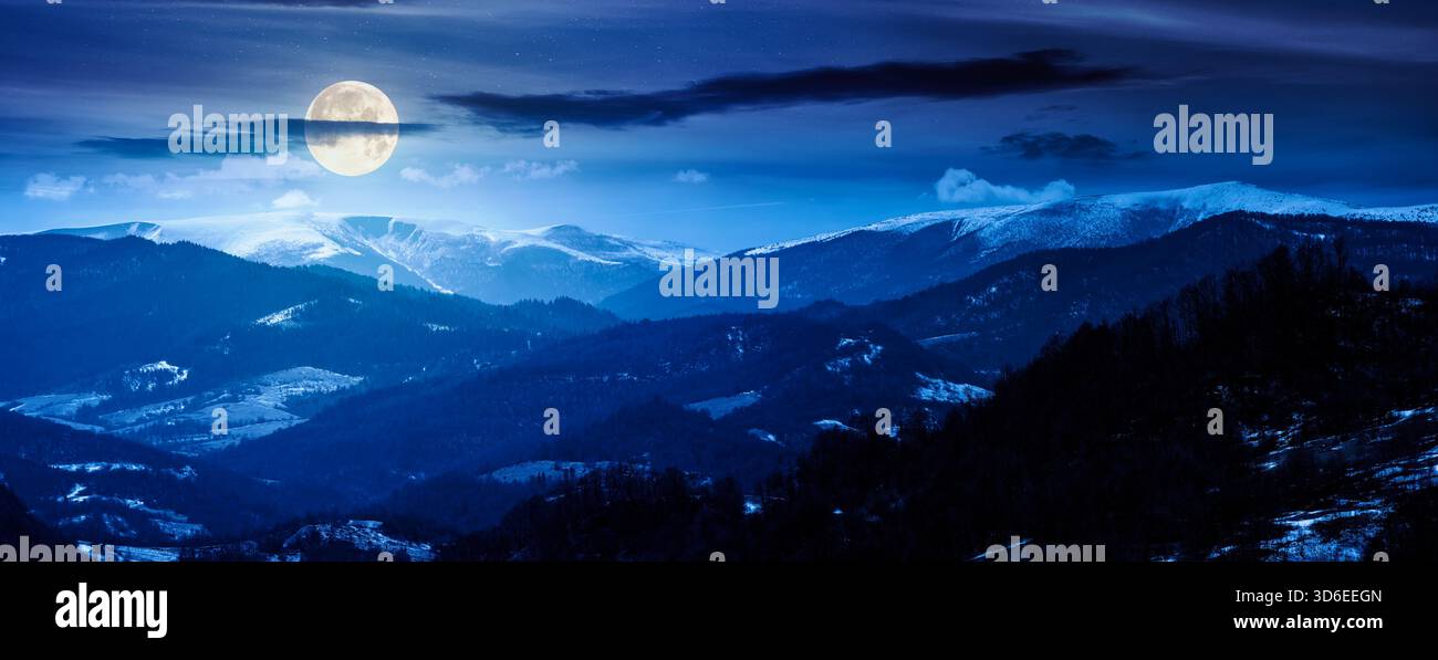 panorama of rolling hills and mountain ridge with snow cowered tops at night. beautiful landscape in springtime cold weather in full moon light. backg Stock Photo