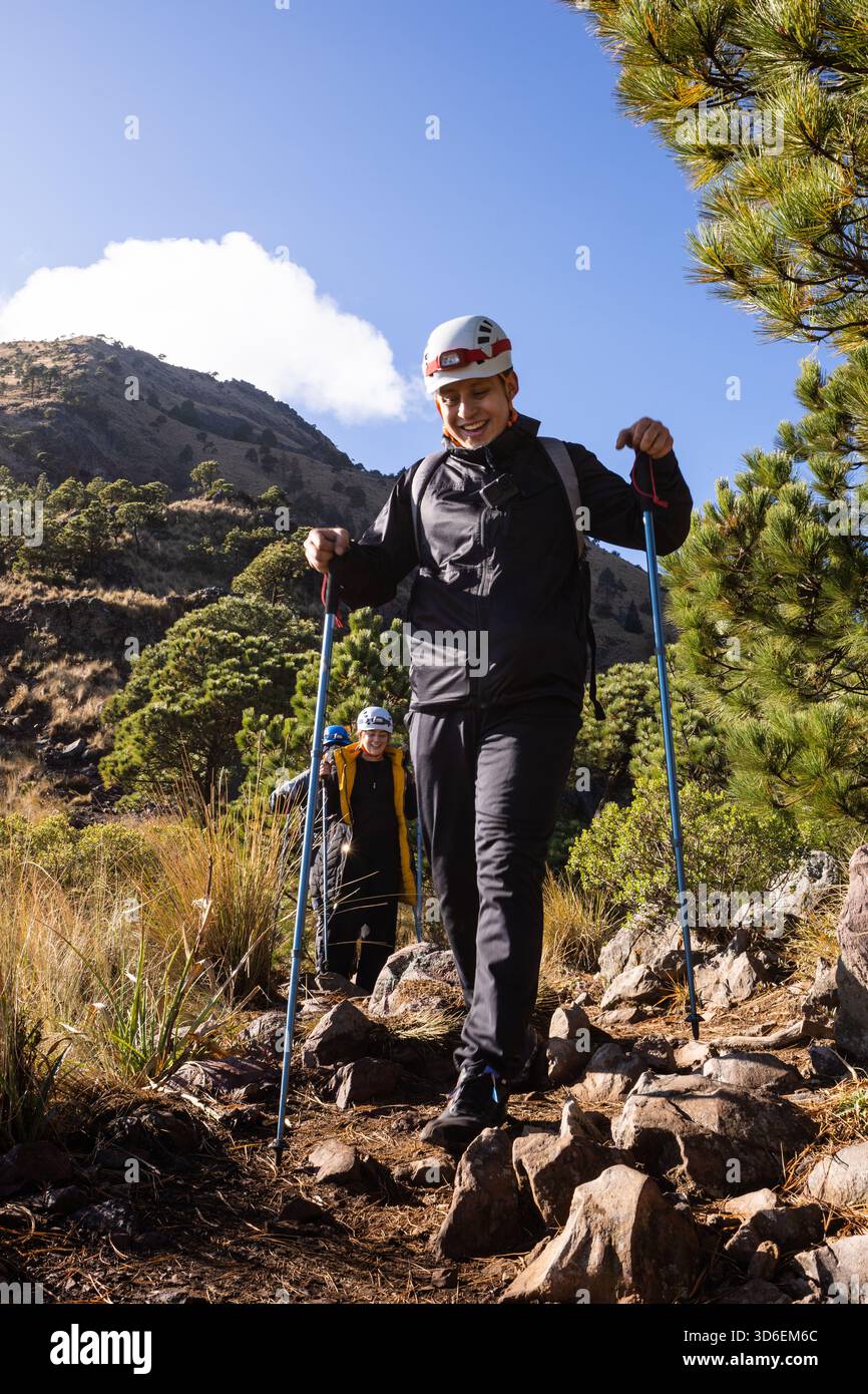 Enthusiastic hikers navigate rugged trails in sunny weather, wearing helmets and using poles. Framed by green foliage, they embody adventure and the g Stock Photo