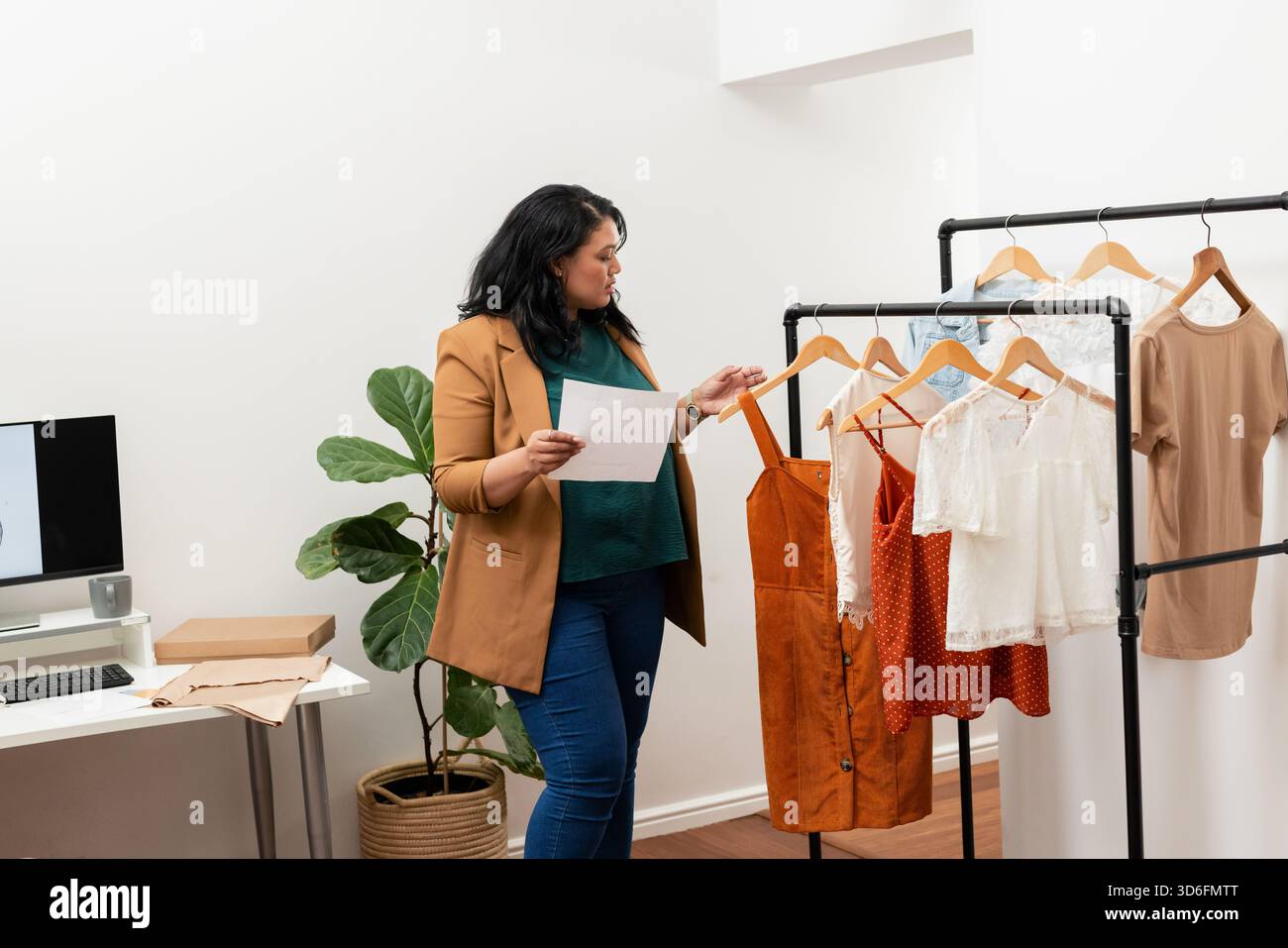Asian woman holding paper while browsing black metal clothing rack with wooden hangers in office Stock Photo