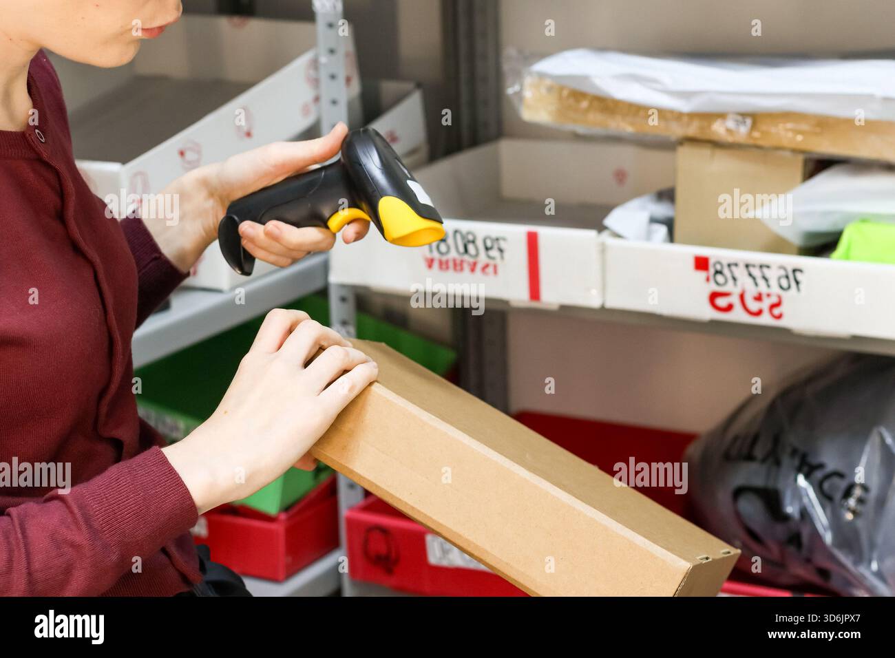 Female worker using handheld scanner on box label while holding package in storage rack Stock Photo