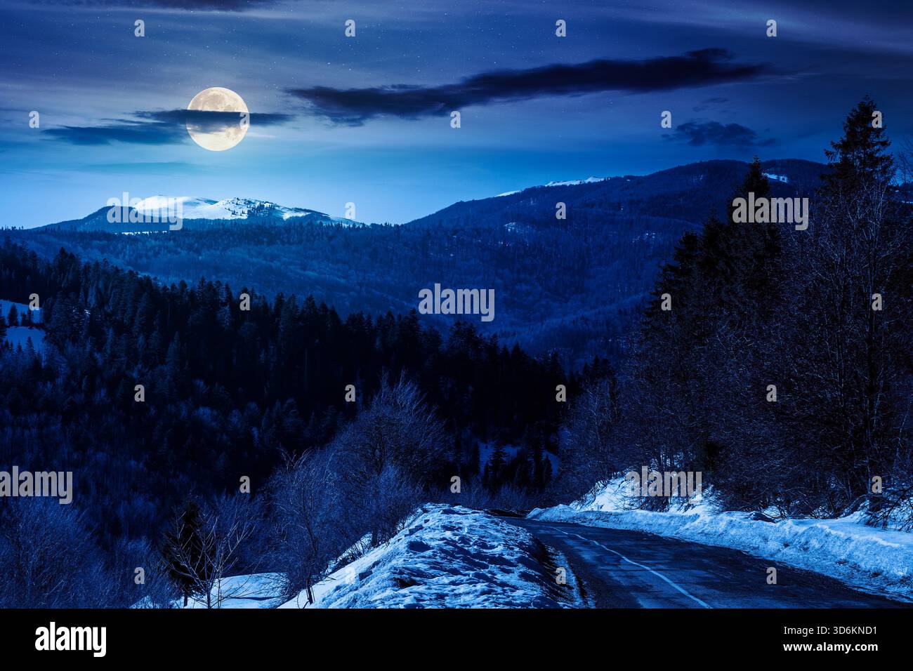 country road winding through winter landscape under dark sky at night. mountains in full moon light. travel background with pass and snow covered slop Stock Photo