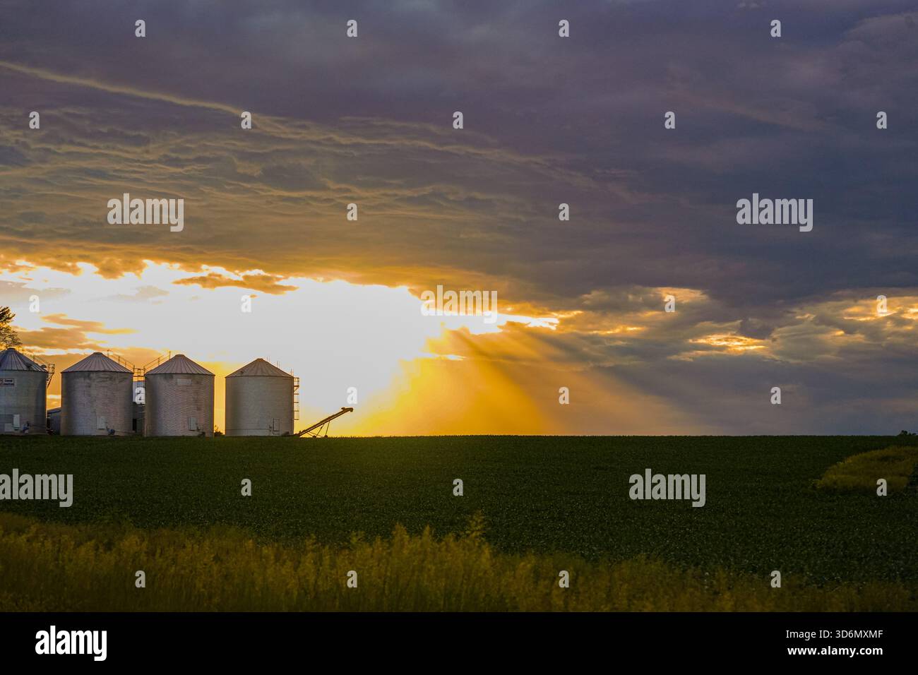Sun Rays Shining Through Clouds Behind Grain Bins Stock Photo