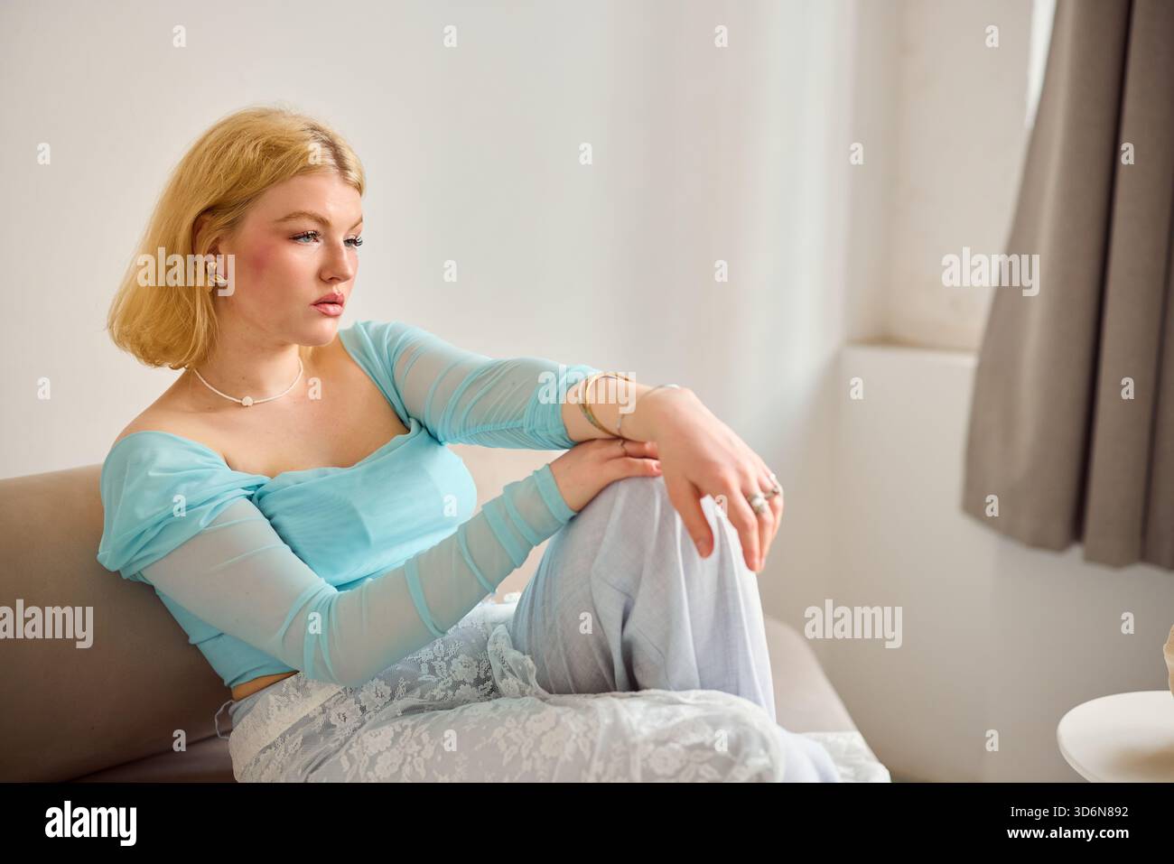 Young woman in light blue top sitting on sofa, looking pensive Stock Photo