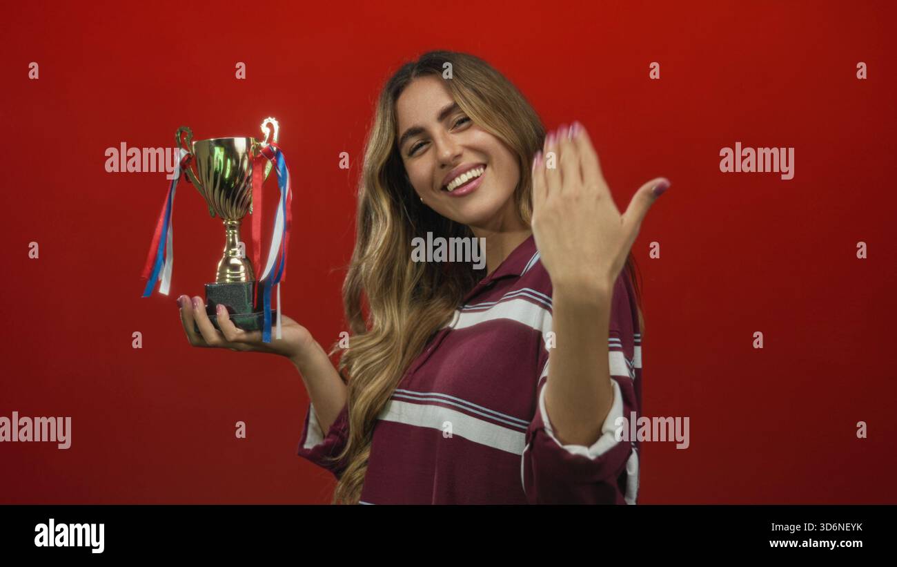Woman holds trophy in studio with finger pointing to trophy highlighting the cup against red background; achievement celebration success pride. Stock Photo