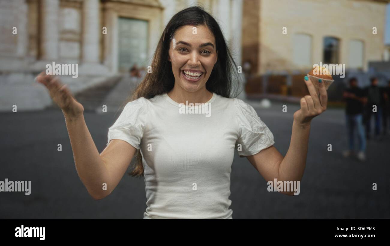 Young hispanic woman beckons with hand while holding muffin on street near historic building, smiling to camera; joy friendly invite. Stock Photo