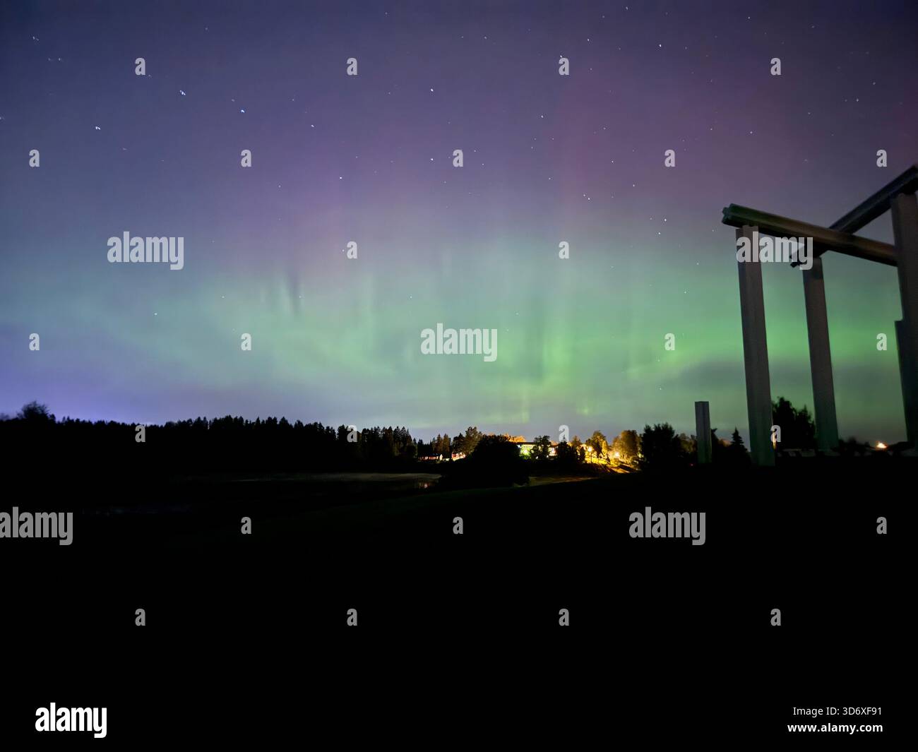 Green and purple auroras shine above open fields and distant trees, casting a soft glow across the peaceful night. - Smartphone Captured Stock Image