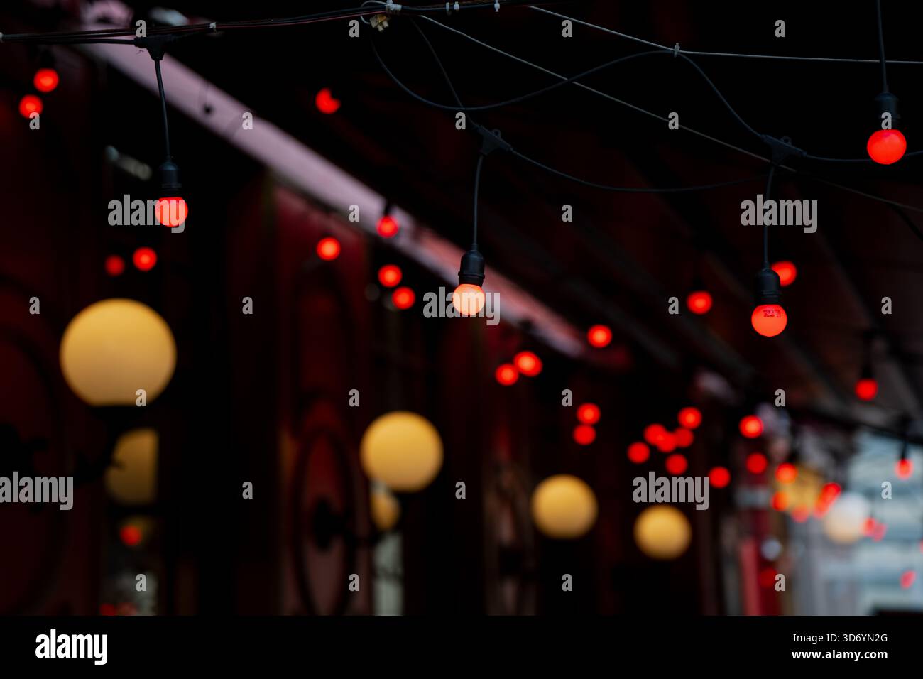 Hanging string lights with glowing red bulbs suspended on dark cables above decorative wall lamps in an atmospheric outdoor passage creating layered i Stock Photo