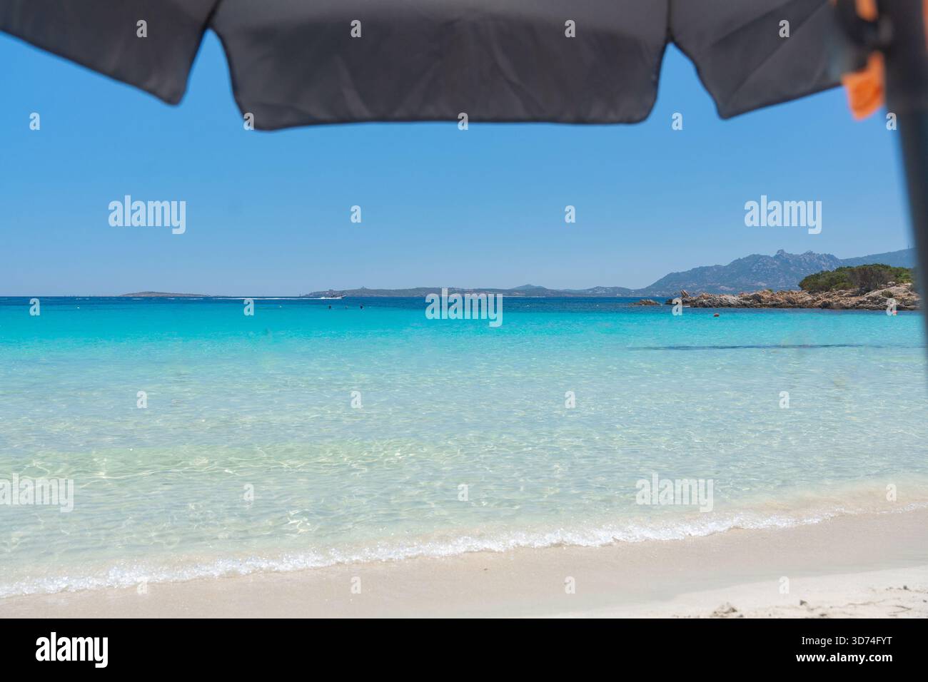 A bright beach view framed by the shade of an umbrella. Clear shallow water and a distant coastline create a peaceful holiday setting with a sense of Stock Photo