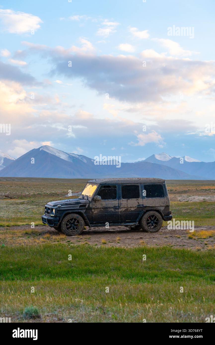 Naryn, Kyrgyzstan - May 13, 2024: Mercedes-Benz G-Class G-Wagon SUV, heavily splattered with mud, parked on a grassy plain with snow-covered mountains Stock Photo
