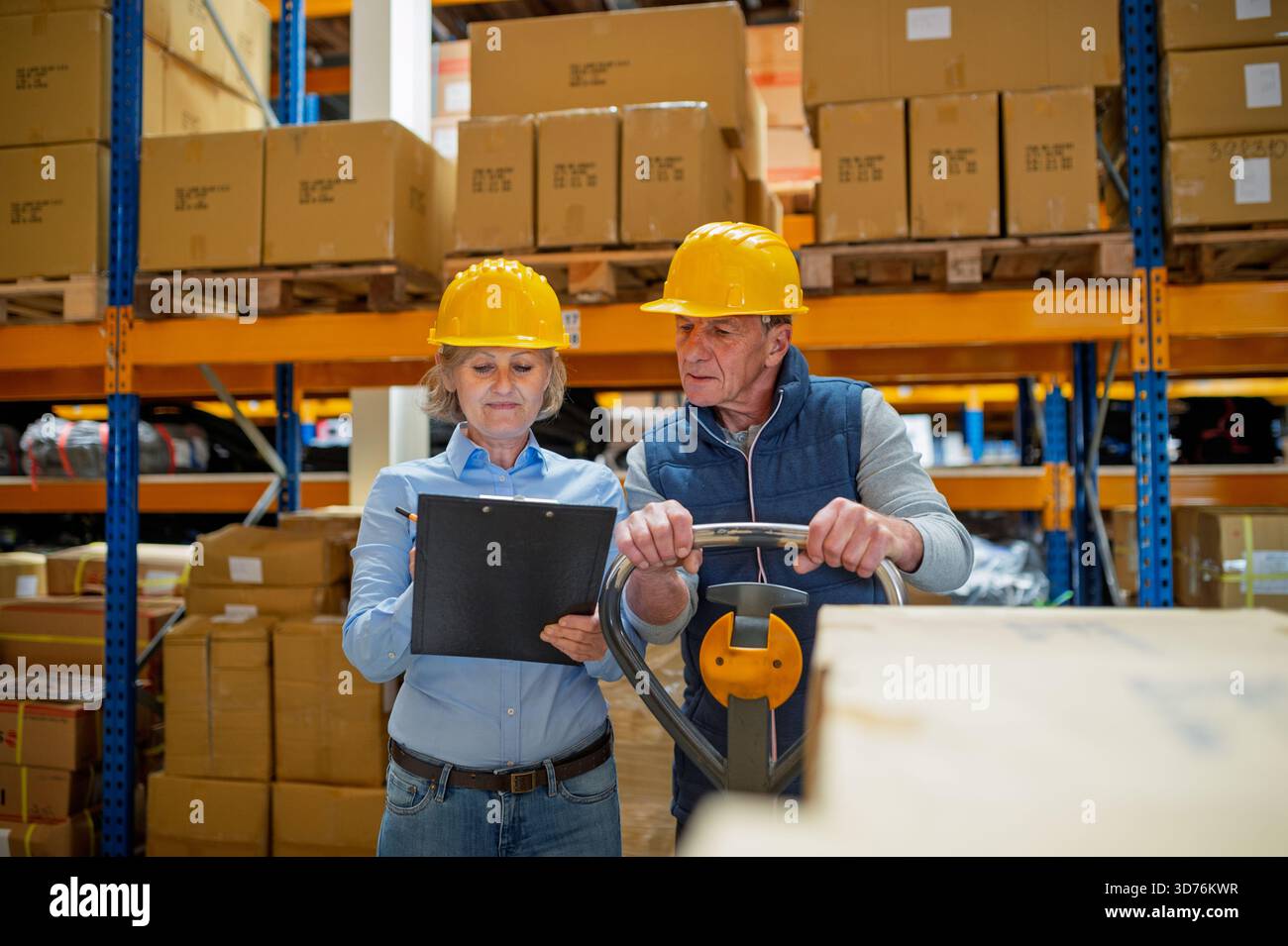 Older manager talking with worker in a warehouse, reading plan document. Stock Photo