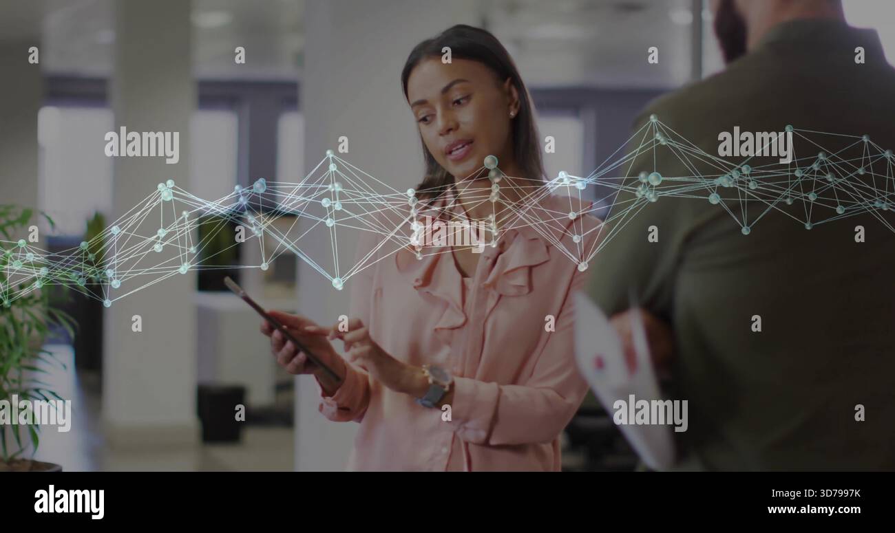 Pointing woman in pink blouse holding tablet and speaking in office, graph overlay, papers Stock Photo