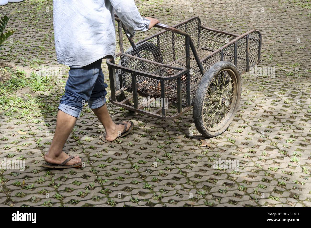 Asian worker pushing metal cart for manual labor on paved outdoor path ...