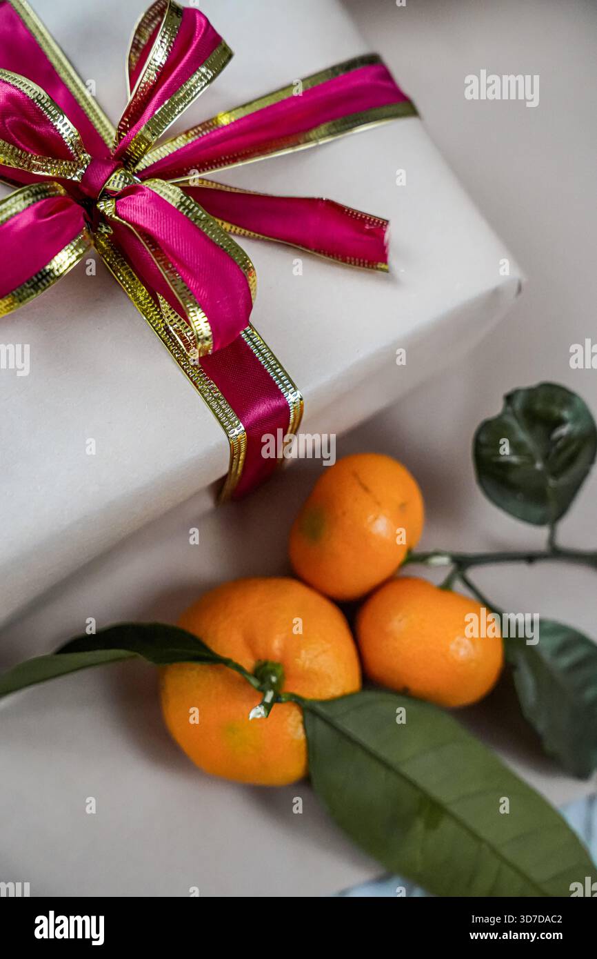 Close-up of a beige gift box decorated with a red ribbon, accompanied by two fresh tangerines on a neutral background. Minimal and festive composition Stock Photo