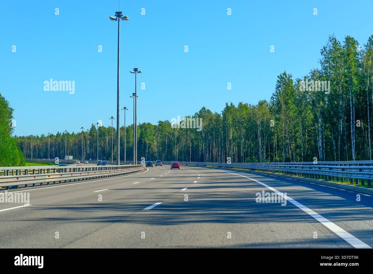Wide multilane highway with street lights outside the city surrounded by deciduous forest on a sunny morning. Stock Photo