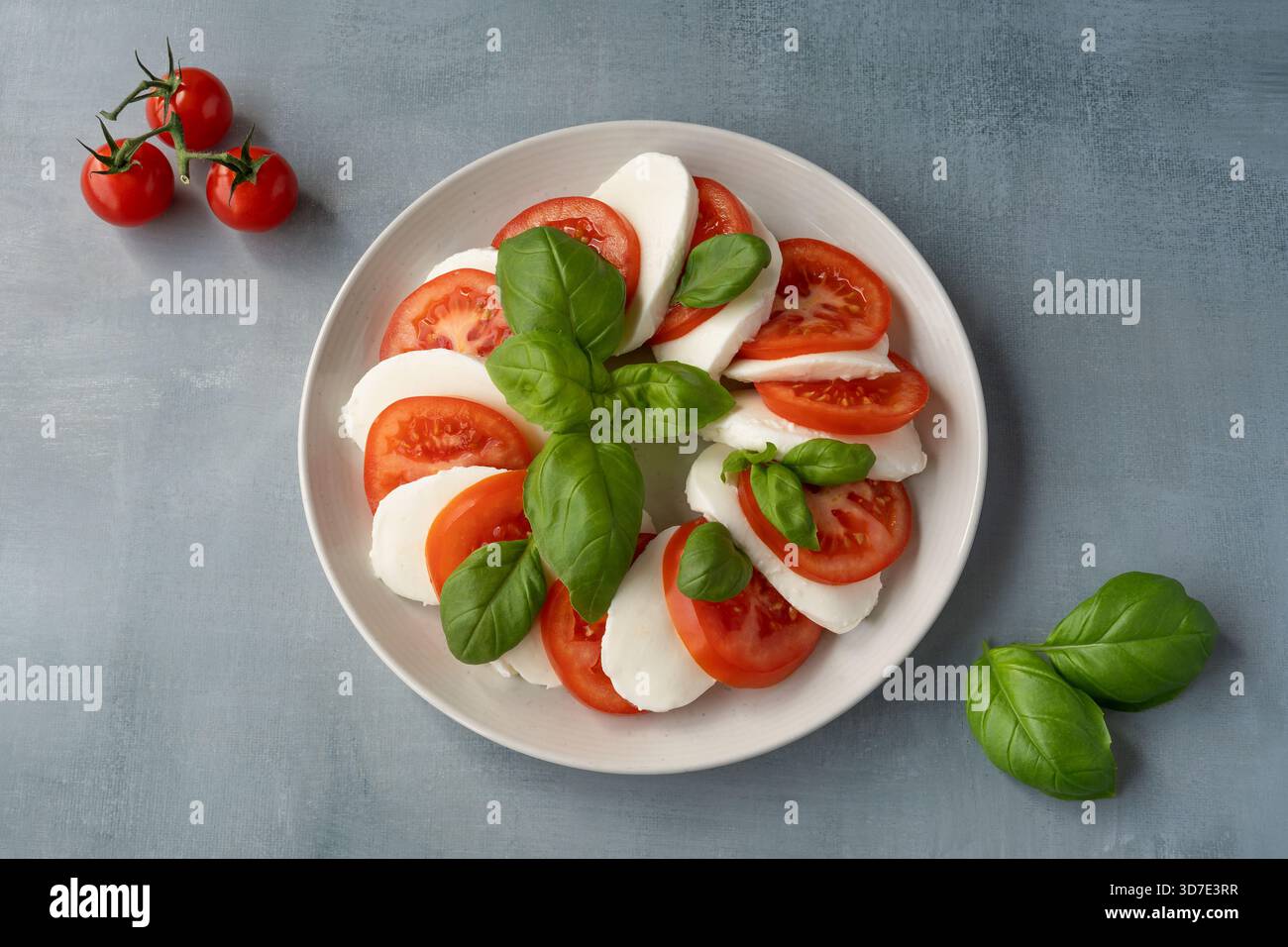 Classic Caprese salad with sliced tomatoes, mozzarella, and basil leaves on a white plate against a blue background. Stock Photo