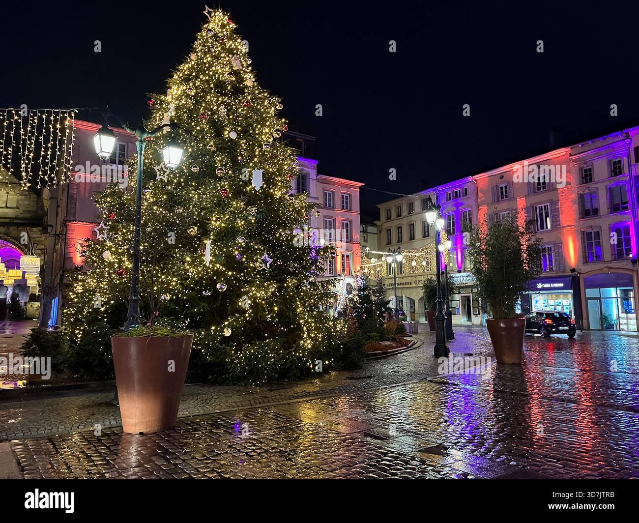 Épinal, France – 2 December 2024: A festive holiday tree illuminated with warm lights stands in the center of the square, surrounded by decorated buil - Smartphone Captured Stock Image