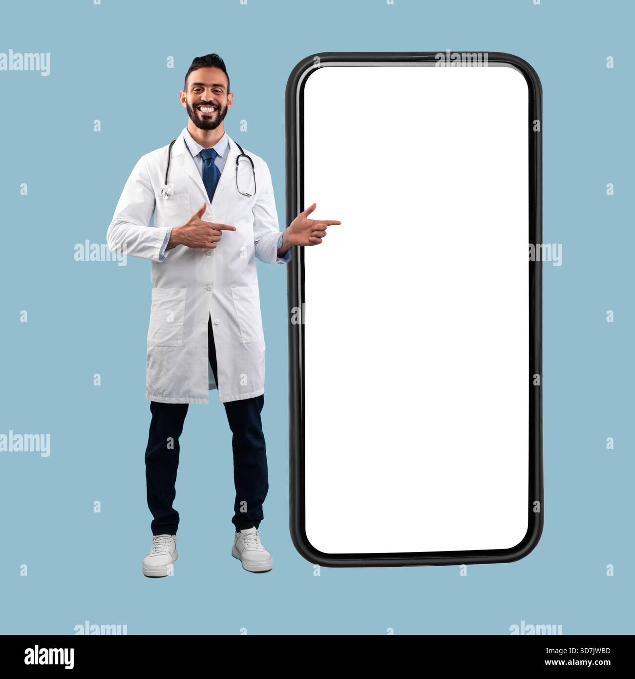 Doctor Standing With a Blank Smartphone Against a Light Blue Background, Pointing With a Smile Stock Photo