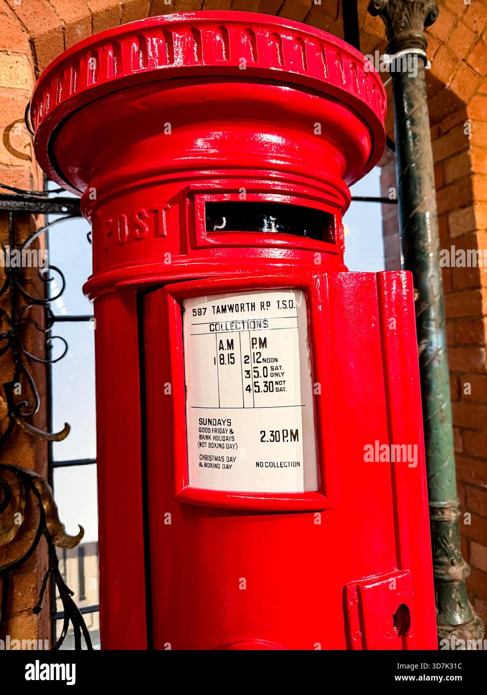 Bright red British victorian VR pillar post box with collection times displayed, against abackdrop of brick architecture & iron window bars - Smartphone Captured Stock Image