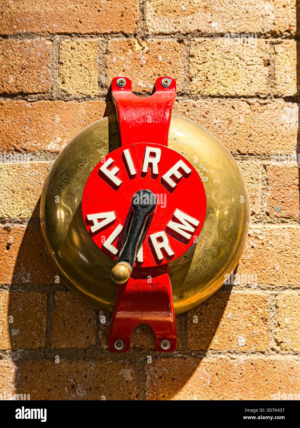 Close-up of a vintage red and brass fire alarm bell mounted on a brick wall under direct sunlight - Smartphone Captured Stock Image