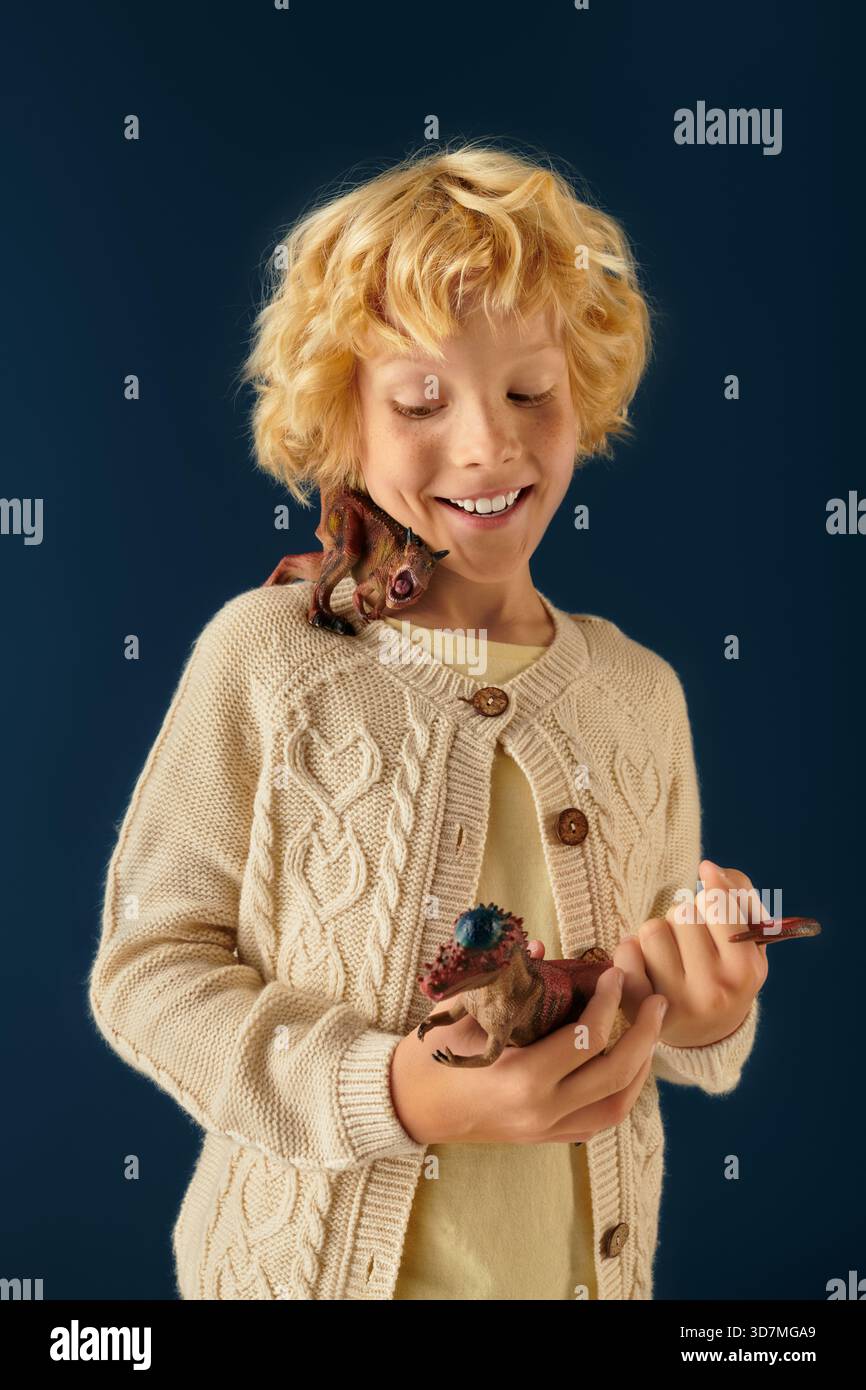 A cheerful blonde boy holds toy dinosaurs in his hands while smiling ...