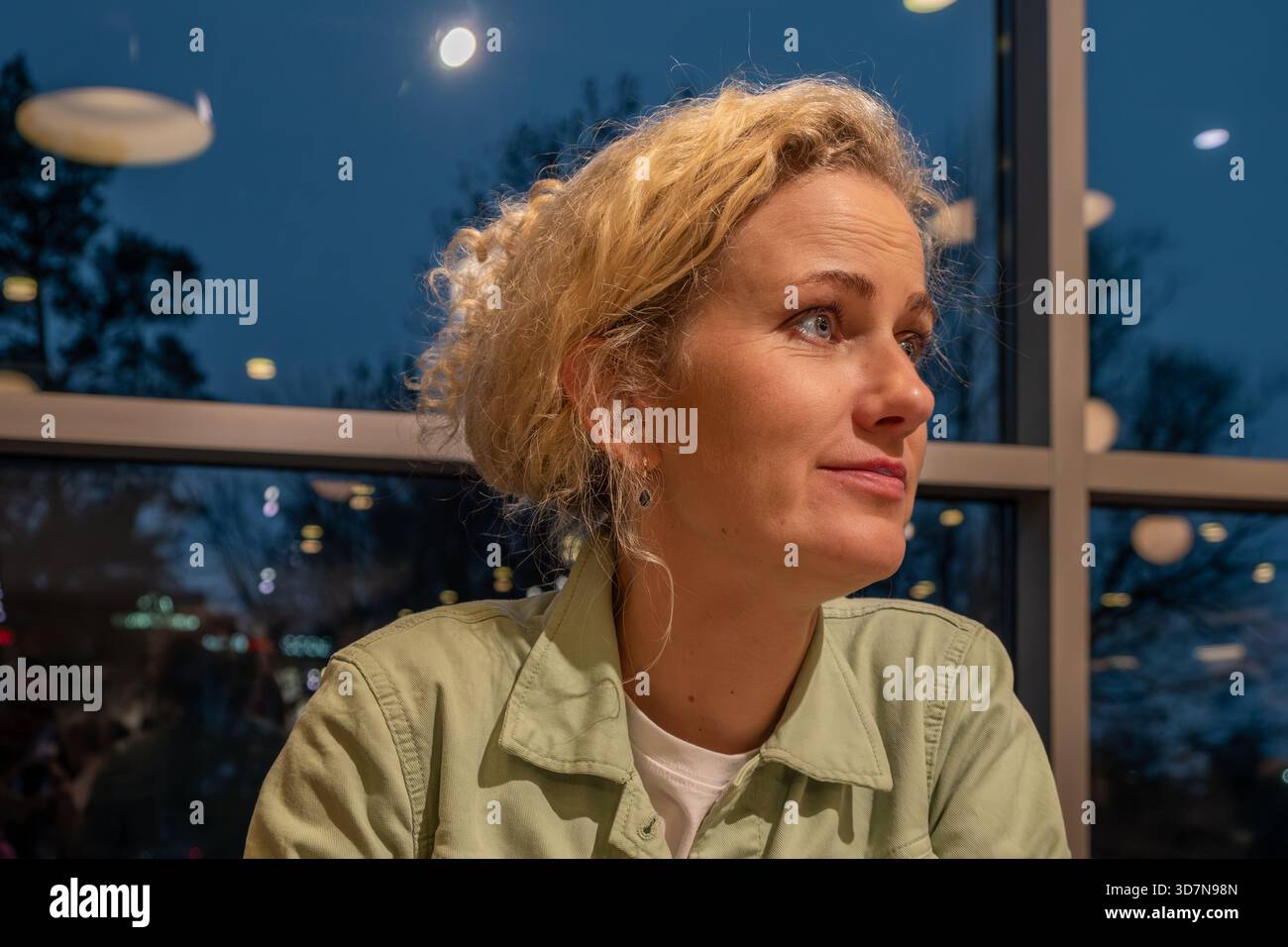 A young woman with curly hair, dressed in a light green jacket, enjoys a serene moment in a cafe, looking Stock Photo