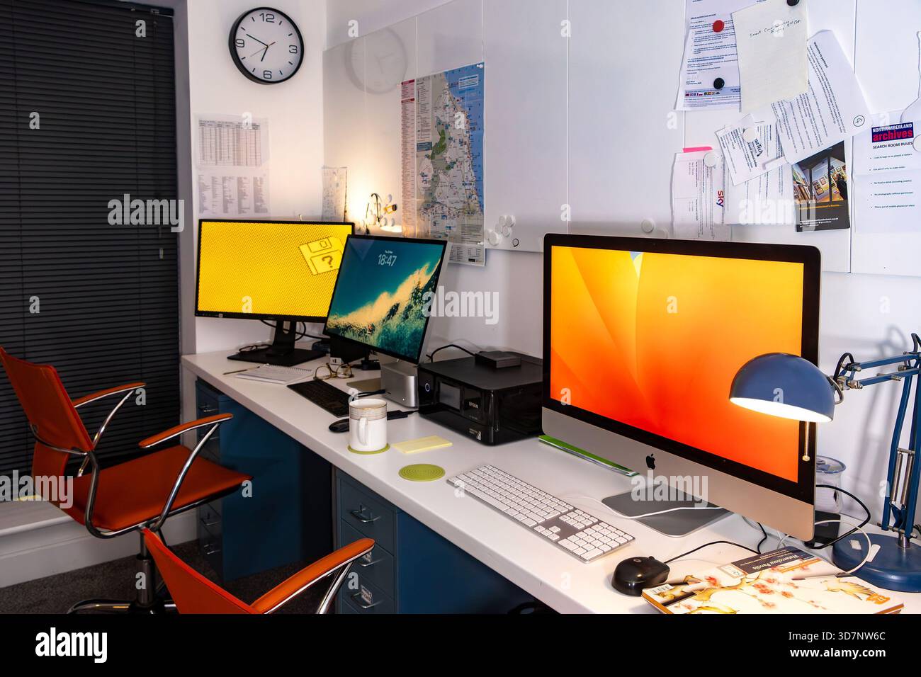 Modern shared office workspace with multiple computer monitors, apple macs, wall clock, documents, and ergonomic chairs in a compact organized setting Stock Photo