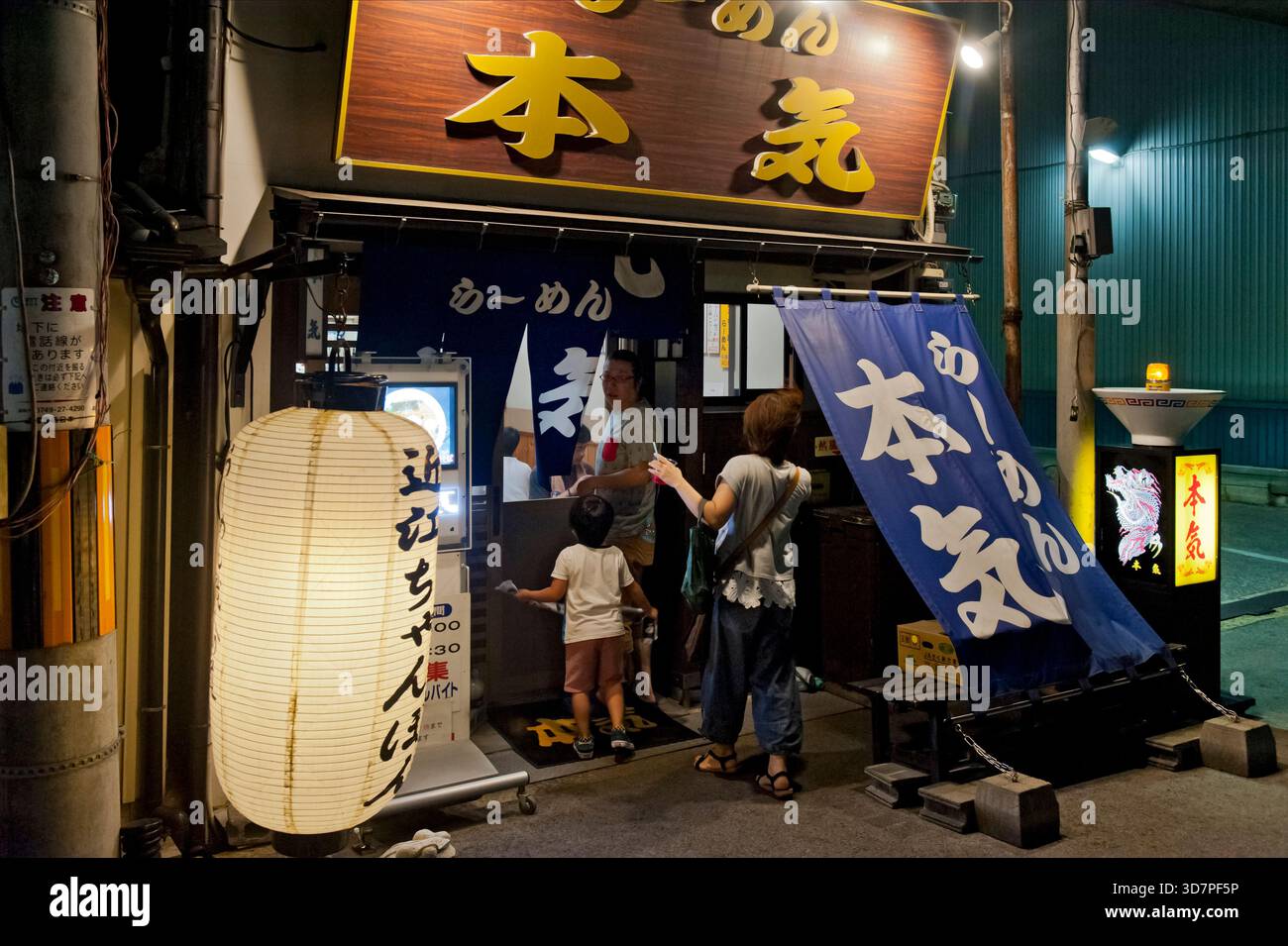 Japanese family entering ramen noodle soup shop "Honki" on a summer night in Hikone, Shiga, Japan. Inviting glowing chochin lantern and noren curtain. Stock Photo