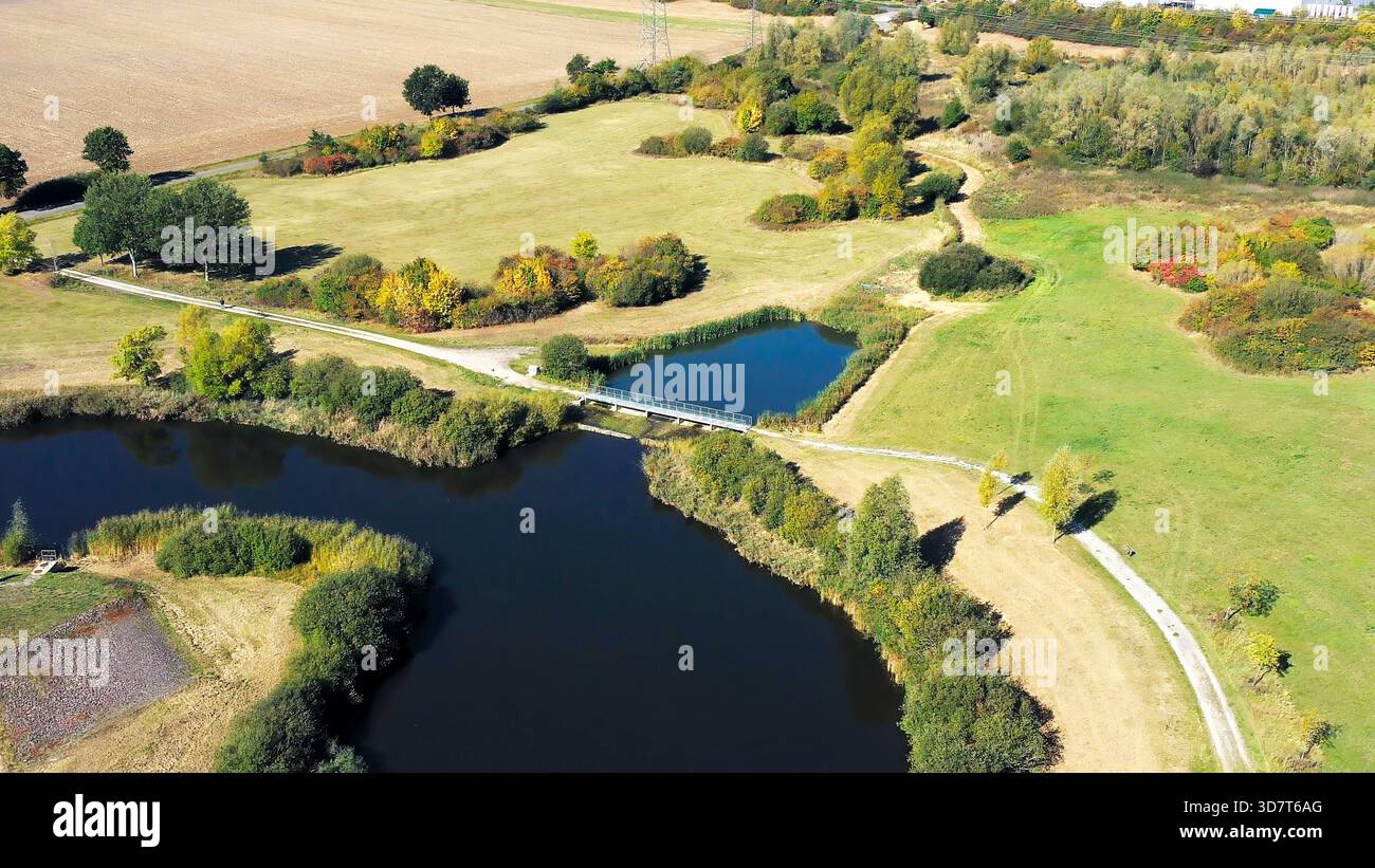 Aerial drone flying above rural nature scene featuring dark lake, connecting pond, footbridge, and fields with changing foliage Stock Photo