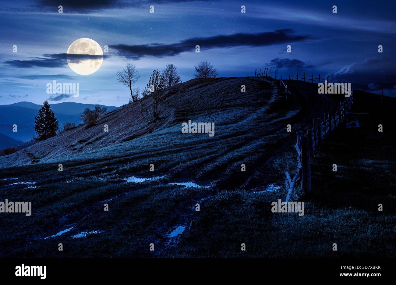 wooden fence near the country dirt road through the grassy hill in spring at night. beautiful nature scenery of mountainous rural area with epic sky a Stock Photo
