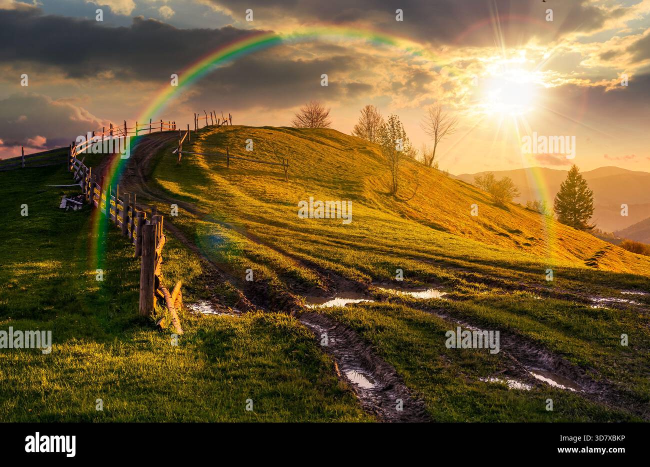 wooden fence near the country dirt road through grassy hill in spring at sunset. beautiful scenery of mountainous rural area in evening light. storyte Stock Photo