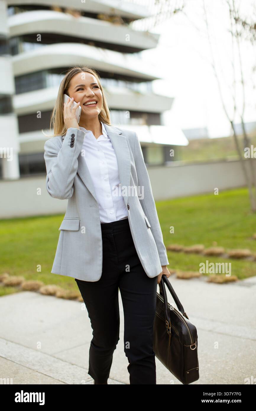 A smiling woman in a light gray blazer walks with confidence while talking on her phone near a contemporary building. The setting features greenery an Stock Photo