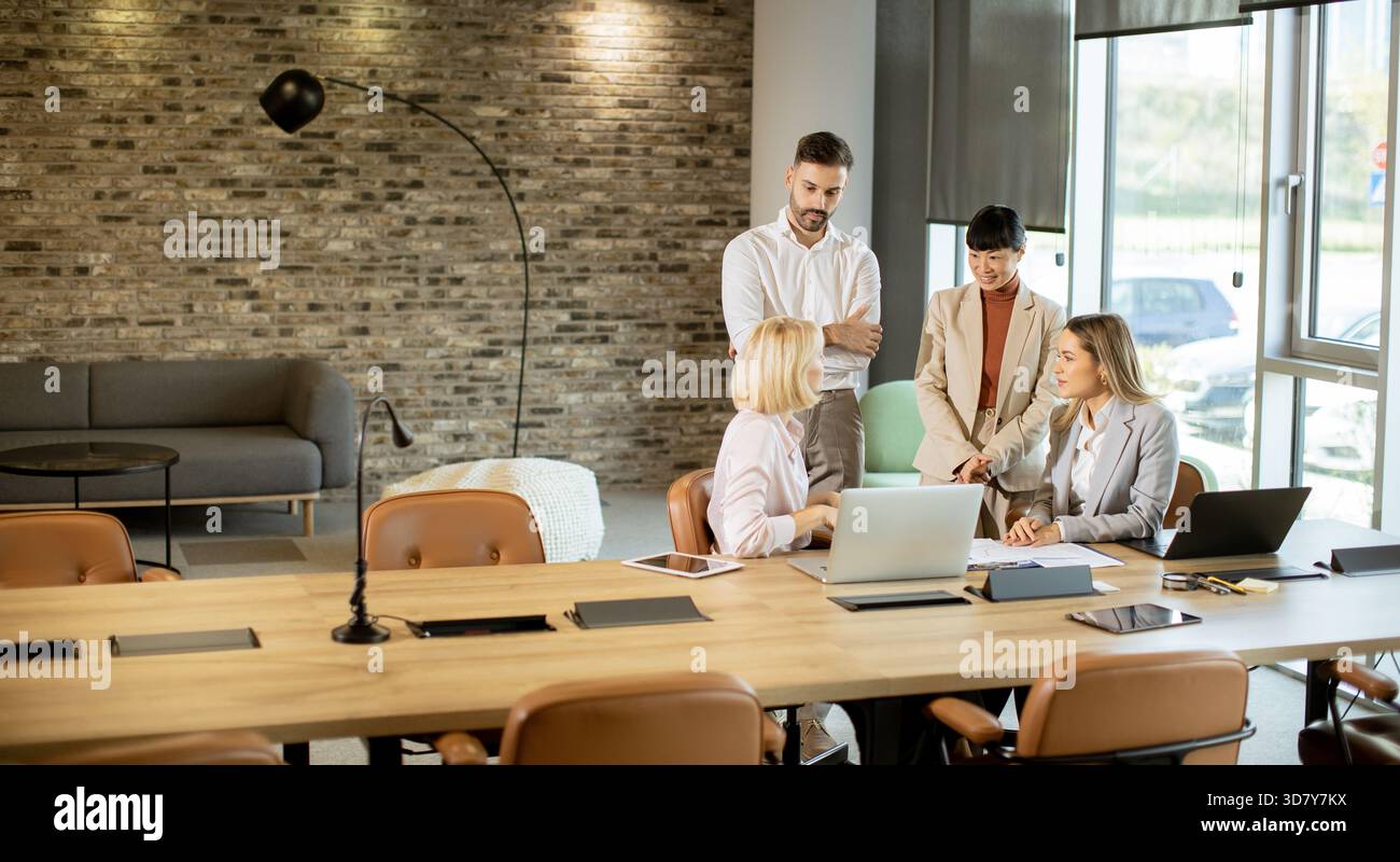 A diverse team engages in a business meeting around a long wooden table. They discuss ideas while laptops and documents are present, creating a collab Stock Photo