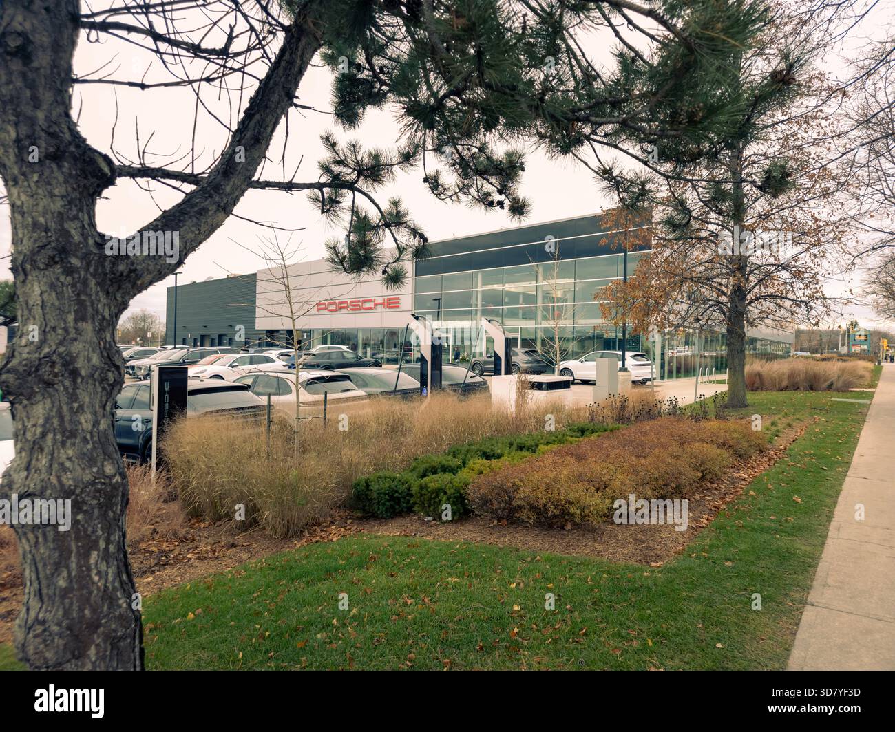 Porsche dealership with glass facade, parked luxury cars, landscaped sidewalk, and sparse trees in autumn. - Smartphone Captured Stock Image